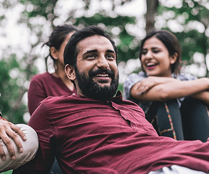 A man in casual clothes with dark hair and a beard lounges outside, smiling at someone off camera.