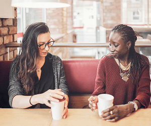 Two women sit side by side with coffee cups speaking