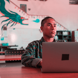 An unsmiling teenaged boy looks up over his laptop in class