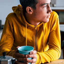 A young man sits at a table holding a mug of coffee and looking thoughtfully to the side