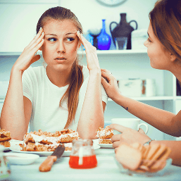 A teenaged girl holds her fingers to her head as she speaks with her mother over breakfast